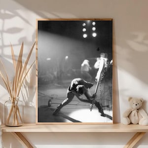 Puede incluir: Fotografía enmarcada en blanco y negro de una actuación musical en vivo. Un músico está inclinado tocando la guitarra en el escenario. El escenario está iluminado con focos y otros miembros de la banda son visibles en el fondo.