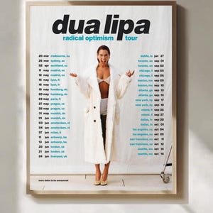 以下が含まれることがあります： デュア・リパの「ラディカル・オプティミズム」ツアーの日程が記載されたフレーム入りポスター。白いバスローブを着たデュア・リパの写真が掲載されています。ツアーの日程は、ポスターの左右の列に記載されています。