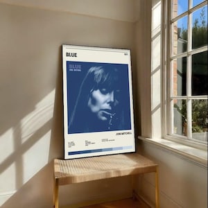 May include: A framed poster featuring the album cover for Joni Mitchell's "Blue" album. The poster has a blue and white color scheme, with a portrait of Mitchell. The poster is displayed on a small, woven bench with gold legs.