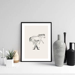May include: Black framed art print with a black and white illustration of a person holding a spotted horse. The artwork is set against a cream background. The print is displayed on a white surface with decorative vases and a plant.