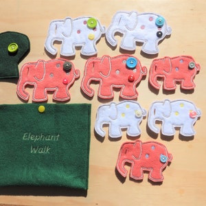 May include: A felt Elephant Walk game set featuring white and coral elephants, each with a button. A green felt tree and a green felt pouch with "Elephant Walk" embroidered on it are also included. The set is displayed on a light wood surface.