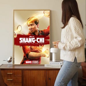 May include: Framed movie poster for "Shang-Chi and the Legend of the Ten Rings" featuring the Marvel Studios logo. The poster has a red banner with the movie title and a central image of the main character. The poster is displayed on a wooden cabinet.