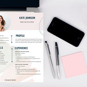 May include: A web developer's CV for Kate Johnson, with a photo, contact details, skills, and experience. The document is on a white surface with a laptop, a smartphone, pens, and a pink sticky note.