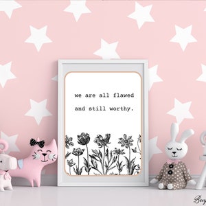 May include: A framed print with the text "we are all flawed and still worthy" above a black and white floral design. The print is displayed with decorative toys on a pink background with white stars.