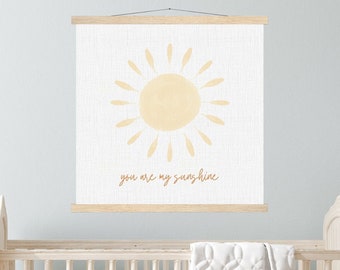 May include: A white crib with a white quilted blanket and a white ruffle skirt. A wooden frame with a white canvas print of a yellow sun with the text "you are my sunshine" hangs above the crib.
