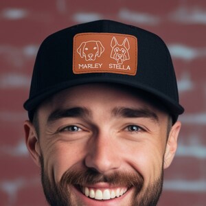 May include: A man wearing a black trucker hat with a brown leather patch featuring two dog silhouettes and the names "Marley" and "Stella". He is smiling and wearing a white t-shirt.