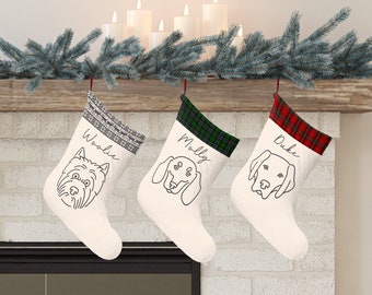 May include: Three white Christmas stockings with personalized names "Woodie", "Molly", and "Duke" and a black and white line drawing of a dog on each stocking. The stockings are hung on a wooden mantel with a garland of pine branches.