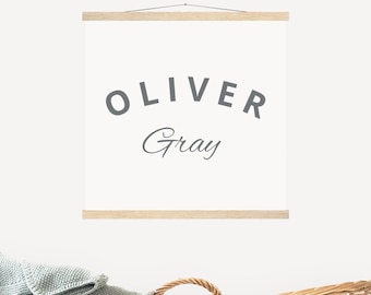 Könnte beinhalten: Ein weißer Leinwanddruck mit dem Namen "Oliver Gray" in grauer Schrift. Der Druck hängt an einer weißen Wand über einem Weidenkorb.