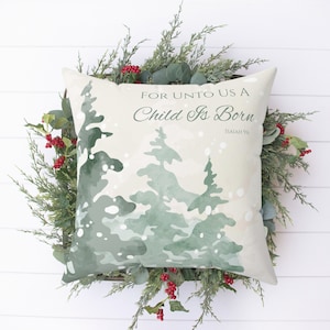 May include: A white throw pillow with a green watercolor design of pine trees and snowflakes. The pillow has the text "For unto us a child is born" and "Isaiah 9:6" printed on it.
