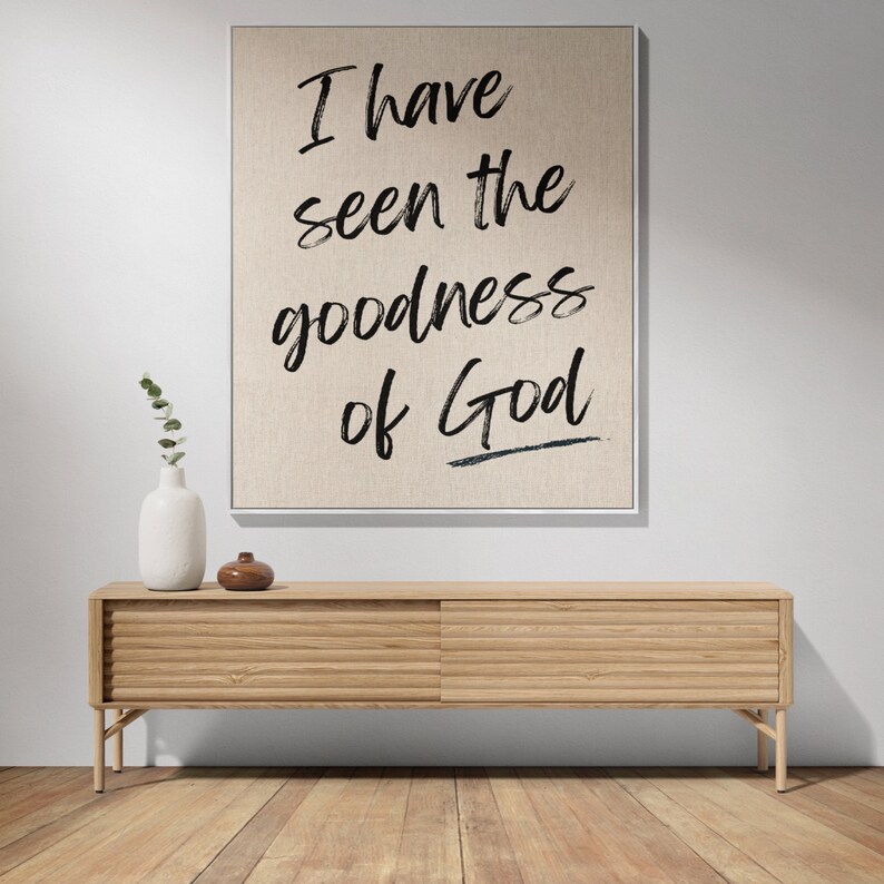 May include: A canvas print with the text "I have seen the goodness of God" in black brush script on a beige background. The print is hanging above a light wood cabinet with two doors.