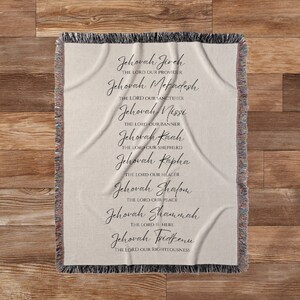 K&ouml;nnte beinhalten: Eine beige gewebte Wurfadecke mit schwarzem Text, der die Namen Gottes auflistet. Die Namen sind in einer dekorativen Schrift geschrieben. Die Decke hat einen Fransenrand und wird auf einer Holzoberfl&auml;che pr&auml;sentiert.