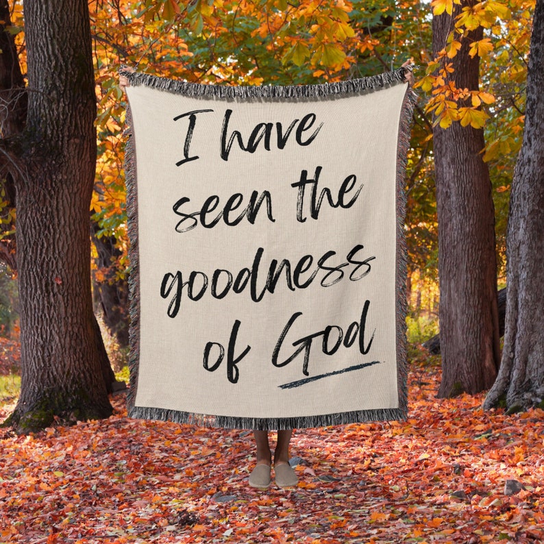 May include: A woven throw blanket with the text "I have seen the goodness of God" in black lettering on a beige background. The blanket is being held up in a woodland setting with autumn foliage.