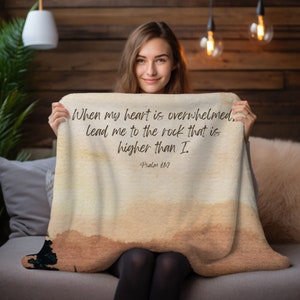 May include: A beige throw blanket with a watercolour background and the text "When my heart is overwhelmed, lead me to the rock that is higher than I. Psalm 61:2" printed in brown.