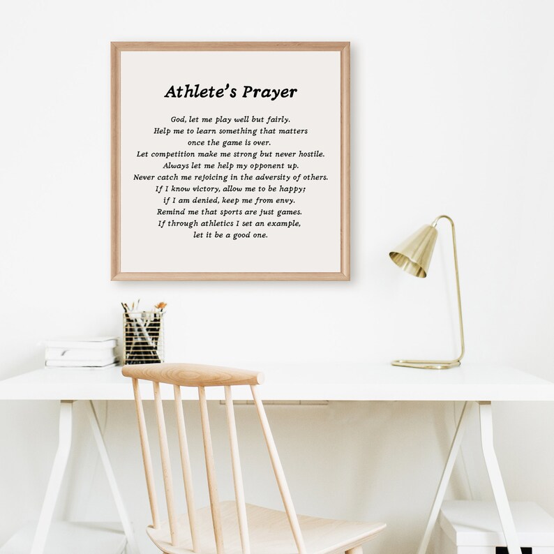 May include: A framed print with a white background and black text. The text is a poem titled "Athlete's Prayer" and includes the following lines: "God, let me play well but fairly. Help me to learn something that matters once the game is over." The print is on a white desk with a gold lamp and a wooden chair.