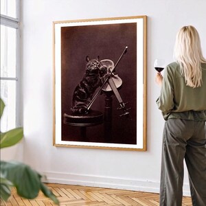 May include: A sepia-toned framed print featuring a cat playing a violin. The cat, with dark tabby markings, stands on a small stool, holding the bow. The violin is mounted on a stand. The print is in a wooden frame.
