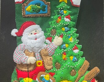 Op de afbeelding: Een groene kerstsok van vilt met een kerstmanfiguur die een hengel vasthoudt. De sok is versierd met een groene kerstboom van vilt, versierd met pailletten en kralen, een vis in een lijst en een lantaarn.