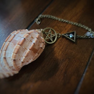 May include: A seashell pendulum with a gold chain and two charms: a pentagram and a triangle with a crystal in the center.