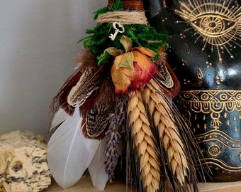 May include: A small, handmade broom with a brown handle, tied with twine and decorated with dried flowers, feathers, and wheat stalks. The broom is resting on a wooden surface with a stone next to it.