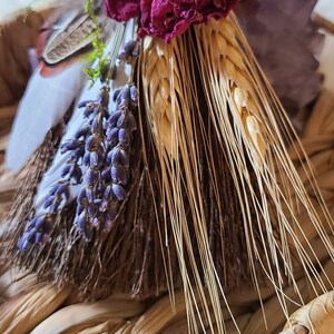 May include: A small, handmade broom with a brown handle wrapped in twine. The broom is decorated with dried lavender, wheat, a dried rose, and a small silver charm. The broom is sitting in a woven basket.
