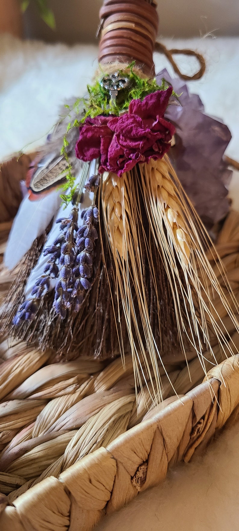 May include: A small, handmade broom with a brown handle wrapped in twine. The broom is decorated with dried lavender, wheat, a dried rose, and a small silver charm. The broom is sitting in a woven basket.