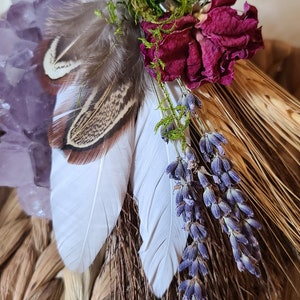 May include: A handmade broom with a brown leather handle, decorated with feathers, dried lavender, a dried rose, and a crystal. The broom is tied with twine and has a key charm.