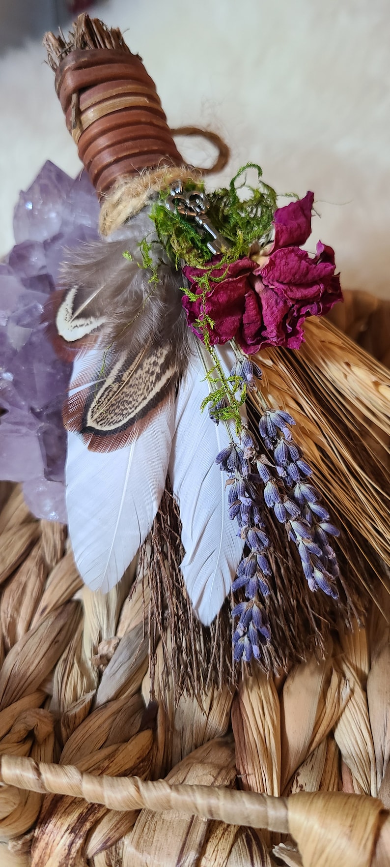 May include: A handmade broom with a brown leather handle, decorated with feathers, dried lavender, a dried rose, and a crystal. The broom is tied with twine and has a key charm.