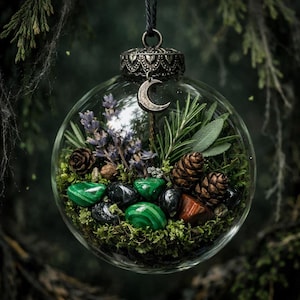 Puede incluir: Adorno esférico de cristal transparente lleno de musgo verde, piedras verdes y negras, piñas, lavanda y romero. Un dije de media luna plateada cuelga de la parte superior.