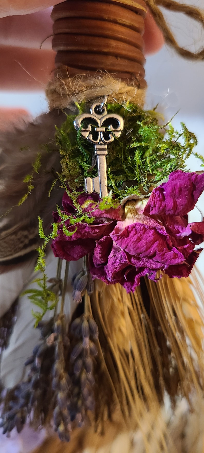 May include: A small broom with a wooden handle wrapped in twine and decorated with dried flowers, moss, and a silver key charm.