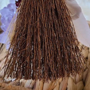 May include: A small brown broom with a woven handle and a white feather tied to the top. The broom is sitting in a woven basket.
