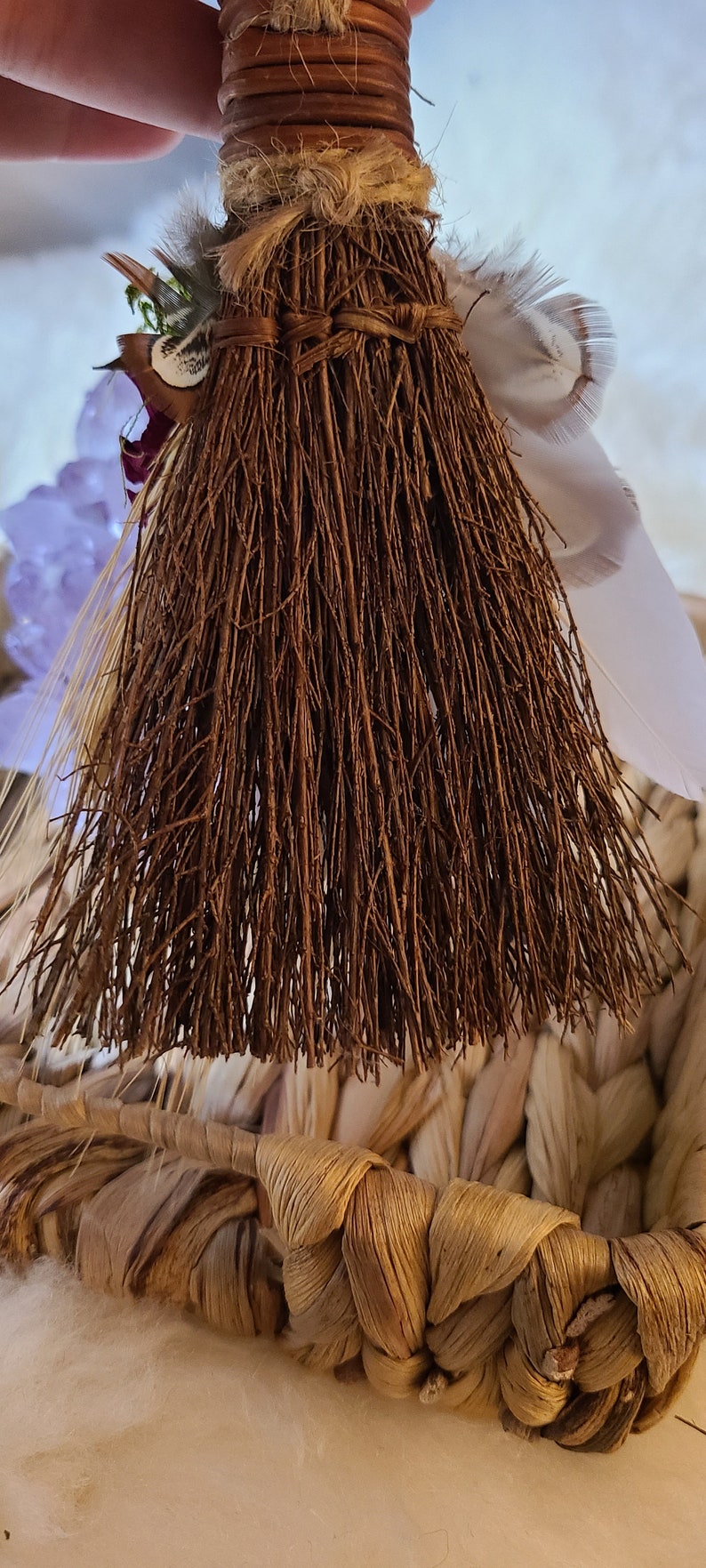 May include: A small brown broom with a woven handle and a white feather tied to the top. The broom is sitting in a woven basket.