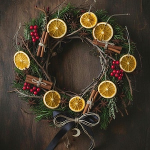 May include: A festive wreath made with dried orange slices, cinnamon sticks, and red berries, set against a backdrop of green foliage. The wreath is decorated with a black ribbon and a crescent moon charm.