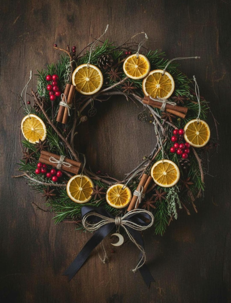 May include: A festive wreath made with dried orange slices, cinnamon sticks, and red berries, set against a backdrop of green foliage. The wreath is decorated with a black ribbon and a crescent moon charm.