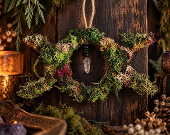 May include: A circular wall hanging crafted from twisted twigs and covered in green, beige, and reddish moss. The structure features three outward-pointing crescent shapes and a central clear quartz crystal pendant suspended by a black bead.