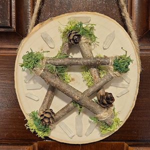May include: A wooden pentagram with five points, made from twigs and tied together with twine. The pentagram is decorated with moss, pine cones, and clear quartz crystals. The pentagram is mounted on a round wooden plaque.