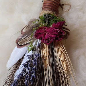 May include: A small, handmade broom with a brown handle wrapped in twine. The broom is decorated with dried lavender, wheat, a dried rose, and feathers. A silver key is attached to the handle.