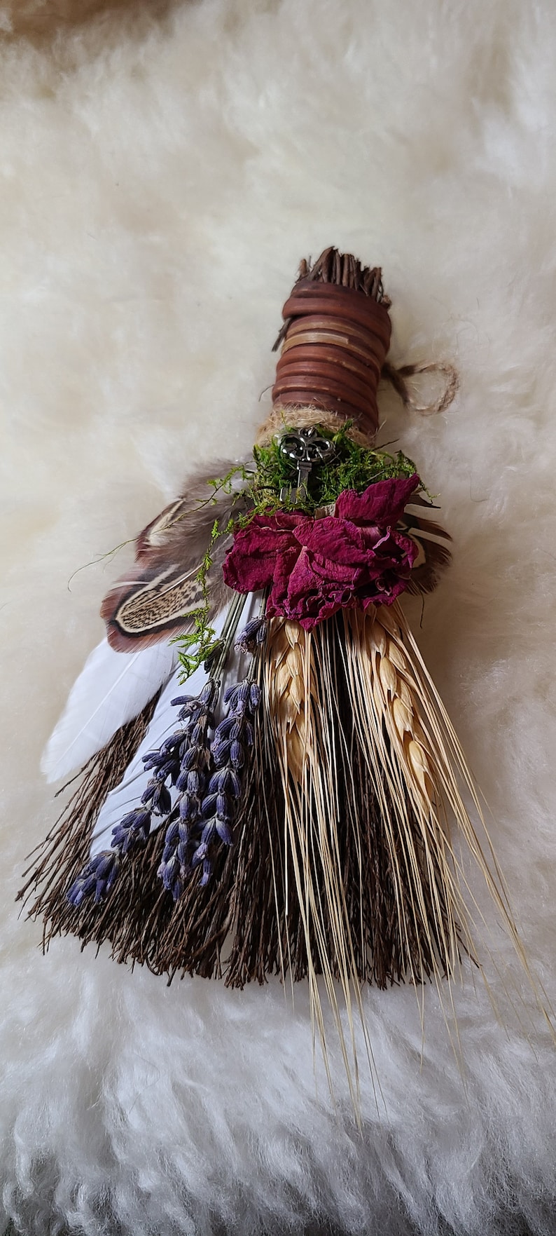 May include: A small, handmade broom with a brown handle wrapped in twine. The broom is decorated with dried lavender, wheat, a dried rose, and feathers. A silver key is attached to the handle.