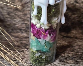 May include: A clear glass jar filled with layers of dried flowers, green moss, and small blue and white stones. The jar is topped with a white wax seal.
