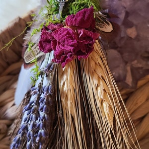 May include: A small, handmade broom with a brown handle wrapped in twine. The broom is decorated with dried flowers, wheat stalks, and a small silver key. The broom is sitting in a woven basket.