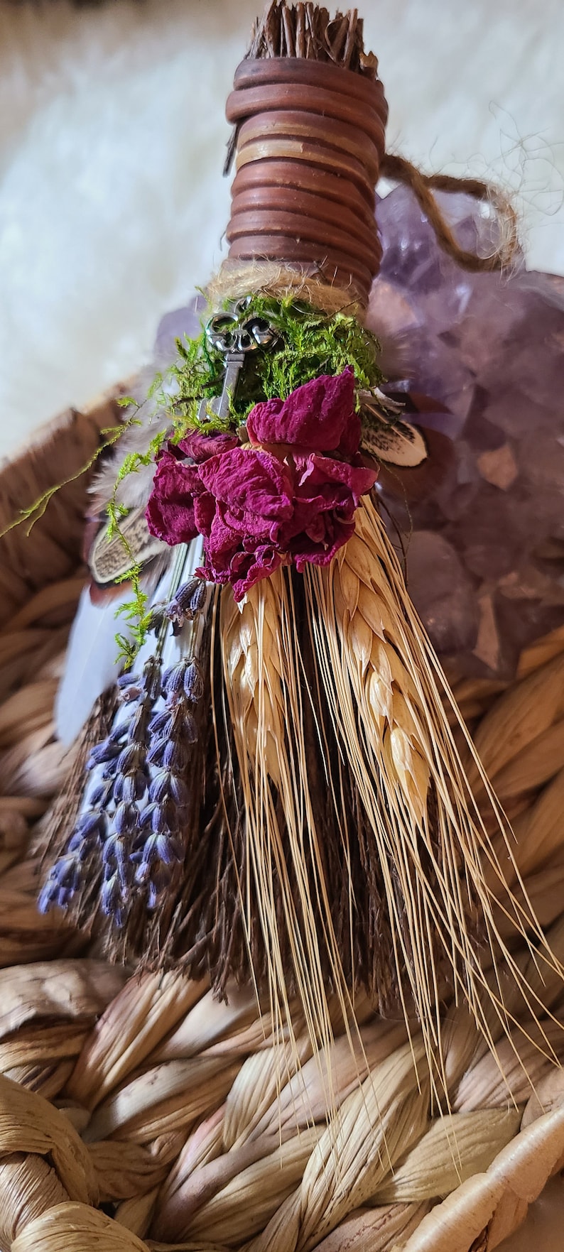 May include: A small, handmade broom with a brown handle wrapped in twine. The broom is decorated with dried flowers, wheat stalks, and a small silver key. The broom is sitting in a woven basket.