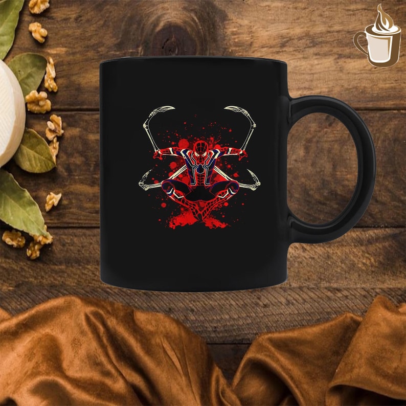 May include: A black ceramic mug featuring a graphic illustration of a red and blue superhero character with four mechanical spider-like appendages extending from the back, set against a background of red paint splatters and dots.