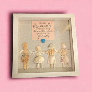 May include: A framed artwork featuring a quote about friends, "It's the friends we meet along the way that help us appreciate the journey." The quote is written on a light brown paper background. The artwork is decorated with four figures made of seashells, holding hands and standing on a beige linen background.