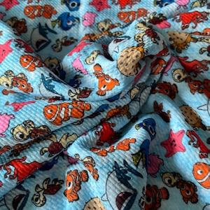 Peut inclure: Tissu bleu avec un motif répété de personnages de Le Monde de Nemo, dont Dory, Nemo, Marlin et d'autres créatures marines. Le tissu a une texture en nid d'abeille.