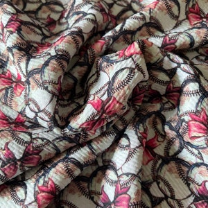 May include: White fabric with a repeating pattern of baseballs and red bows. The fabric has a textured, slightly raised surface.
