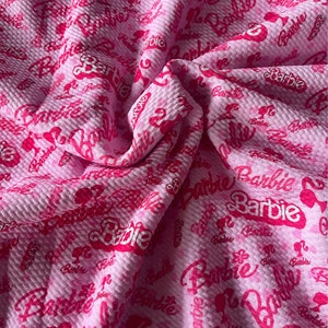 May include: Pink fabric with a textured surface, featuring a repeating pattern of the word "Barbie" in various fonts and styles. The design includes silhouettes of Barbie and floral accents.