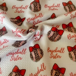 May include: White fabric with a repeating pattern of baseballs with red bows and leopard print. The text "Baseball Sister" is repeated throughout the fabric.