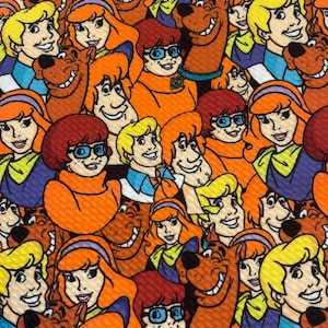 May include: An orange fabric featuring a repeating pattern of cartoon characters, including a brown dog, a blonde-haired man, a woman with orange hair, and a man with red hair and glasses. The characters are smiling and appear to be from a cartoon.