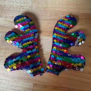 May include: Two sequined, abstract-shaped decorations. Each is covered in colorful, round sequins in shades of blue, green, yellow, red, and silver. The decorations are placed on a wooden surface.