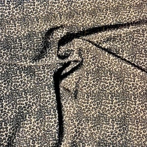 May include: Close-up of a fabric with a leopard print pattern. The fabric has a beige background with black and grey spots. The fabric is folded, creating shadows and texture.
