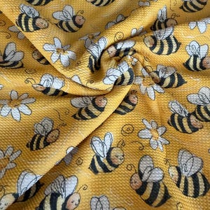 May include: Yellow fabric with a repeating pattern of cartoon bees and white daisies. The bees have black and yellow stripes and white wings. The fabric has a textured surface.