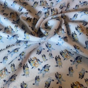 Op de afbeelding: Witte stof met een herhalend patroon van blauwe en gele cartoonhonden. De stof heeft een wafelstructuur.